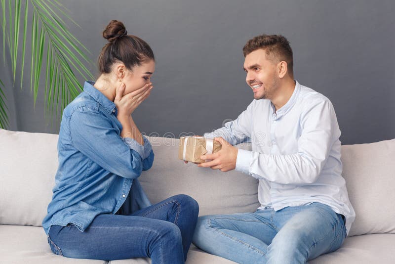 Helgdagskoncept Unga män som ger presenten till en kvinna som täcker det ansikte som är rört på en sofa-studio isolerad på grått. Unga par män som ger en låda till kvinnor som täcker ansiktsleende och som sitter vid sofa studio isolerade på grå bakgrund