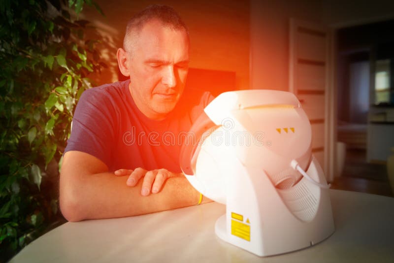 Heling pain with infrared light therapy. Man treating pain in the sinuses and head with infrared light therapy. The man sits at the table near the healing lamp royalty free stock photography