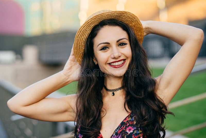 Hembra morena feliz con el sombrero que lleva de la sonrisa agradable que guarda la mano detrás de la cabeza que presenta al aire imagenes de archivo Hembra morena feliz con el sombrero que lleva de la sonrisa agradable que guarda la mano detrás de la cabeza que presenta al aire imagenes de archivo