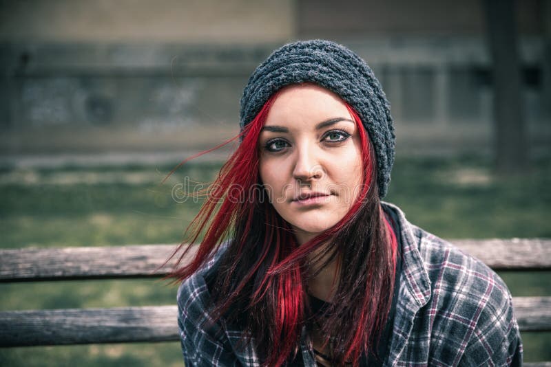 Hemlös flicka, ung härlig röd hårflicka som bara utomhus sitter på träbänken med angelägen hatt- och skjortakänsla och d royaltyfri fotografi Hemlös flicka, ung härlig röd hårflicka som bara utomhus sitter på träbänken med angelägen hatt- och skjortakänsla och d royaltyfri fotografi