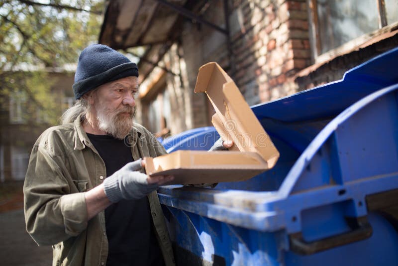 Homeless man standing close to trash can, holding packing from pizza. Tramp looking for food in garbage can. Homeless man standing close to trash can, holding packing from pizza. Tramp looking for food in garbage can.