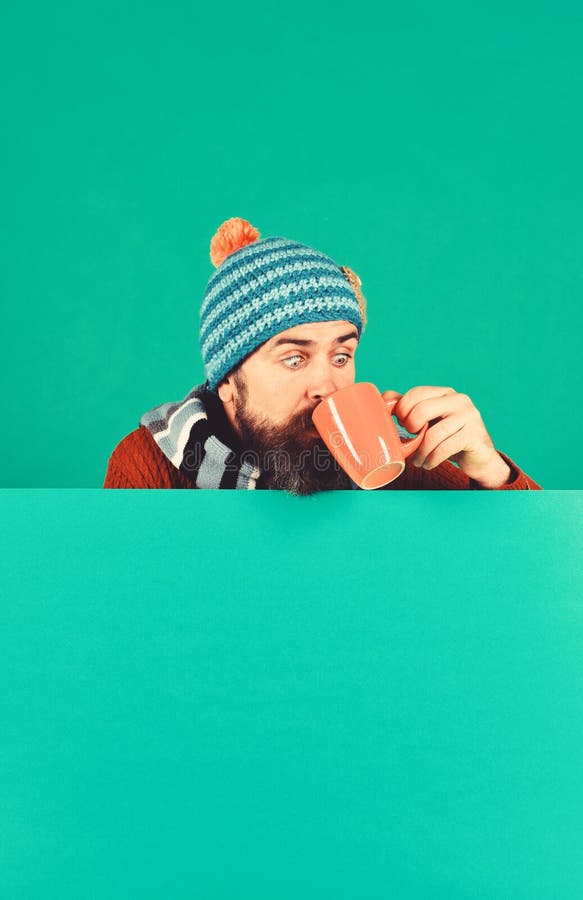 Herbst- und Kältekonzept. Hipster mit Bart und schockiert Gesicht trinkt warmen Tee oder Kaffee. Mann in warmen Hut hält braune Tasse auf grünen Hintergrund Kopierraum. Oktober und November Getränkeidee