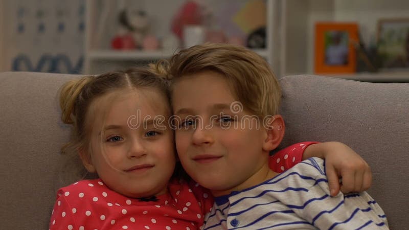 Hermanos felices que abrazan, sentándose en el sofá y mirando la cámara, unidad almacen de video