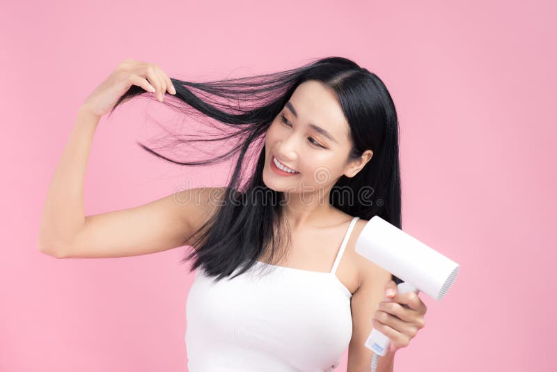 Hermosa Chica Asiática Sonriente Con El Pelo Recto Negro Largo Usando Secador De Pelo Aislado sobre fondo rosa fotografía de archivo libre de regalías