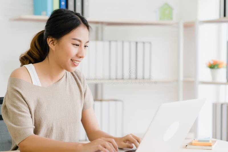 Hermosa joven sonriente asiática que trabaja con una laptop en el escritorio en el salón de casa fotografía de archivo Hermosa joven sonriente asiática que trabaja con una laptop en el escritorio en el salón de casa fotografía de archivo