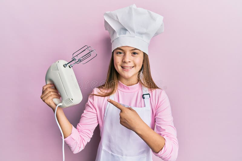 Hermosa morena pequeña niña con sombrero de chef sujetando batidor de bollería mezclador eléctrico sonriente alegre señalando con foto de archivo