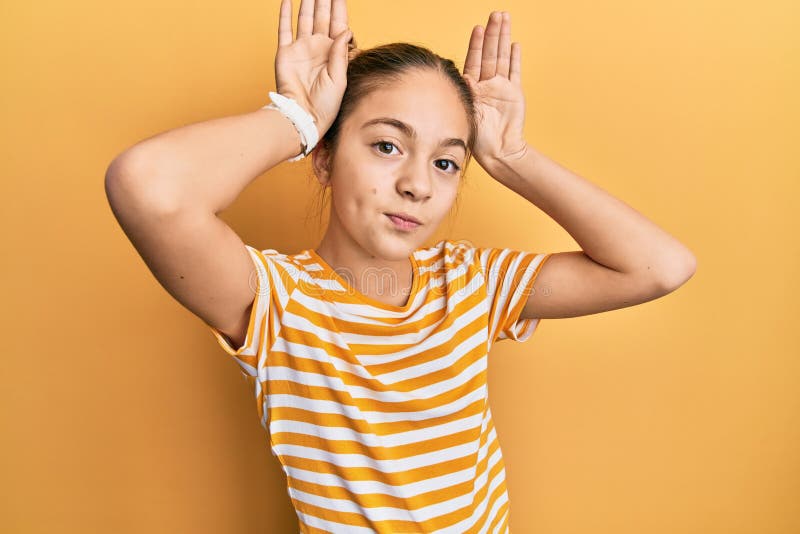 Hermosa morena pequeña niña con una camiseta casual a rayas haciendo gestos de orejas conejitas con las manos palmas de las manos fotografía de archivo