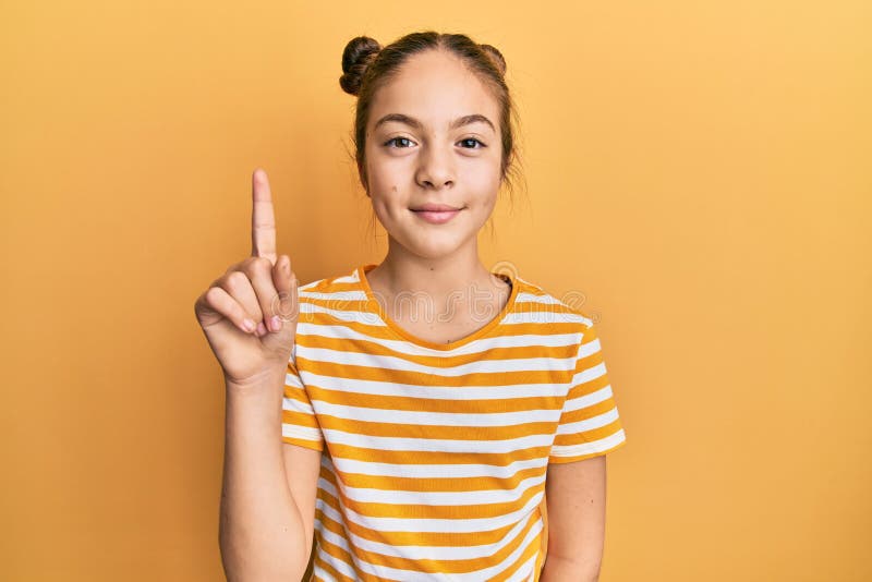 Hermosa morena pequeña niña con una camiseta casual a rayas que aparece y señala con el dedo número uno mientras sonríe foto de archivo
