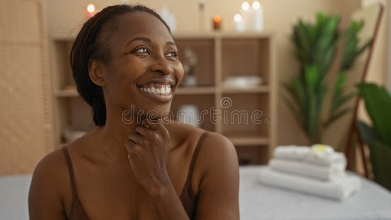 Hermosa mujer africana sonriente en un centro de bienestar con velas y plantas en el fondo, disfrutando de relajación en el interi fotos de archivo