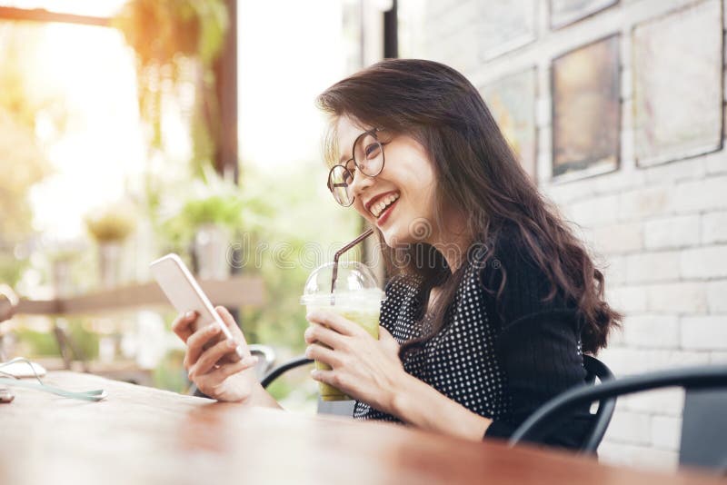 Hermosa mujer asiática más joven bebiendo té verde fresco en una botella y mirando la pantalla de teléfono móvil con cara de foto de archivo