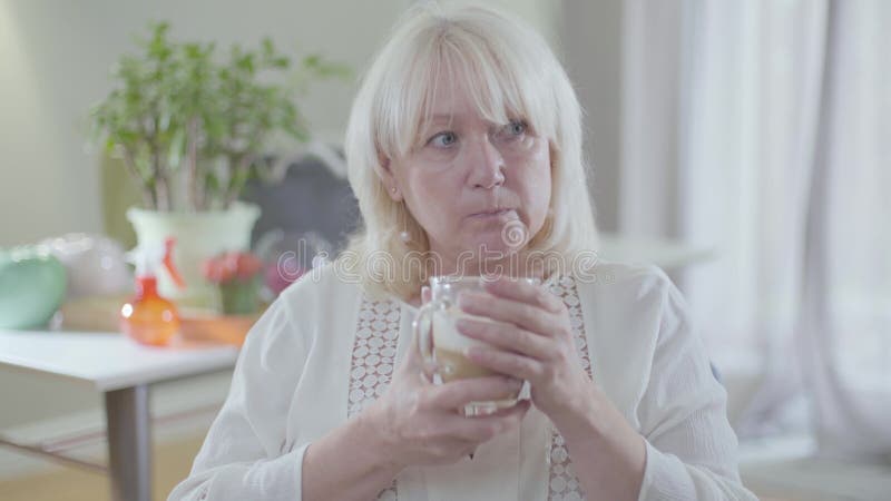 Hermosa mujer caucásica madura con pelo rubio y ojos grises tomando café y mirando lejos Gastos de jubilados solitarios almacen de metraje de vídeo