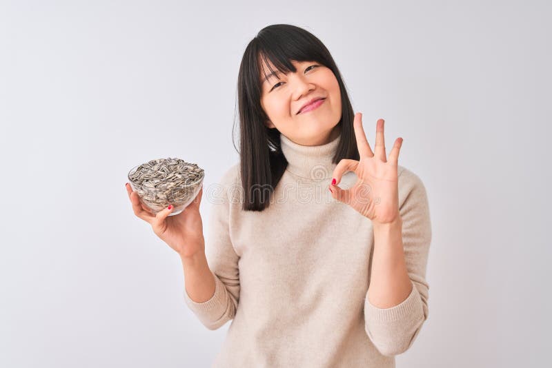 Hermosa mujer china sosteniendo un tazón con semillas de girasol sobre fondo blanco aislado haciendo buen signo con los dedos exce imágenes de archivo libres de regalías