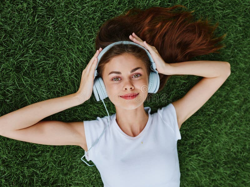 Hermosa mujer con auriculares escuchando música tendida sobre el césped en el parque en primavera y sonriendo con imágenes de archivo libres de regalías