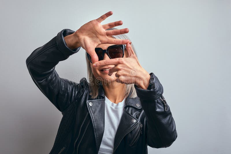 Hermosa mujer con gafas, con una chaqueta de cuero negro sobre un fondo gris, haciendo un marco con las manos con las palmas y fotografía de archivo Hermosa mujer con gafas, con una chaqueta de cuero negro sobre un fondo gris, haciendo un marco con las manos con las palmas y fotografía de archivo