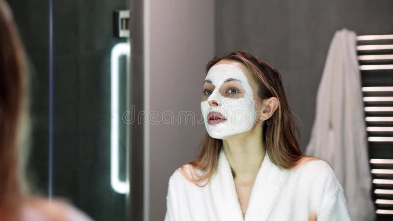 Hermosa mujer en albornoz poniendo máscara blanca para la hidratación usando dedos lento