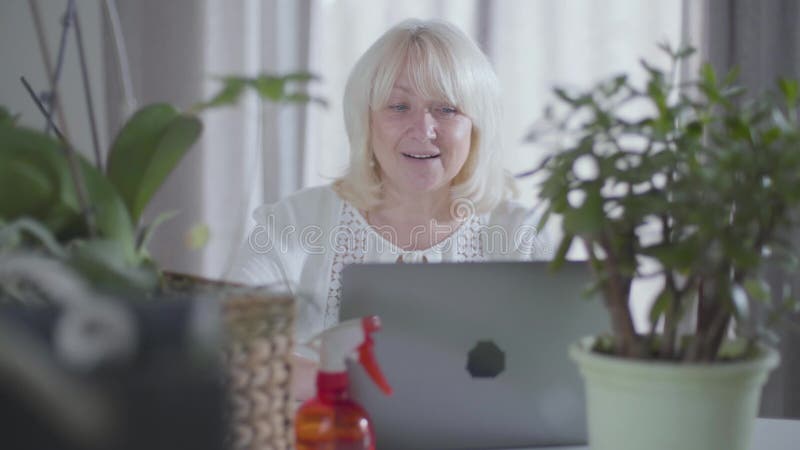 Hermosa mujer rubia y madura sentada con una laptop en casa detrás de su jardinería Retrato de jubilados caucásicos sentados almacen de metraje de vídeo