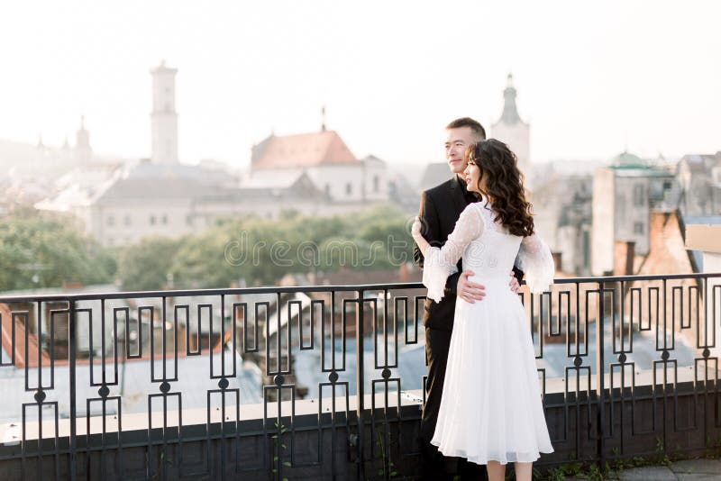 Hermosa pareja de bodas china abrazándose y mirando el amanecer mientras se paran en el fondo de la ciudad vieja fotos de archivo libres de regalías Hermosa pareja de bodas china abrazándose y mirando el amanecer mientras se paran en el fondo de la ciudad vieja fotos de archivo libres de regalías