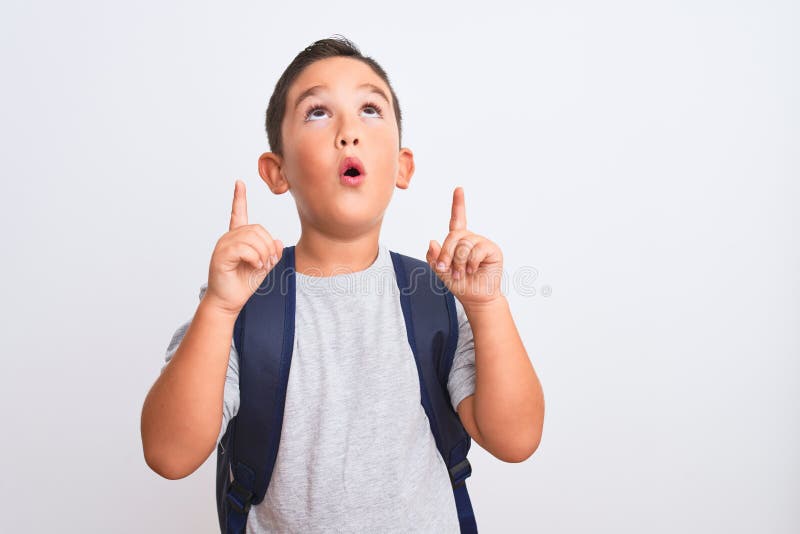 Beautiful student kid boy wearing backpack standing over isolated white background amazed and surprised looking up and pointing with fingers and raised arms. Beautiful student kid boy wearing backpack standing over isolated white background amazed and surprised looking up and pointing with fingers and raised arms