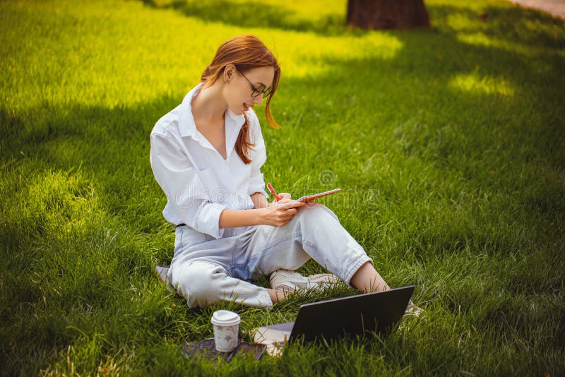 Hermosos estudios de chicas funcionan con el blogger de laptops hace notas de educación a distancia al aire libre en el parque fotos de archivo libres de regalías