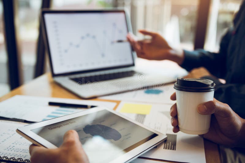 Business partnership pointing to the graph of the company financial statements report and profits earned during in the computer screen with giving a presentation to colleagues holding tablet and coffee in office room. Business partnership pointing to the graph of the company financial statements report and profits earned during in the computer screen with giving a presentation to colleagues holding tablet and coffee in office room.