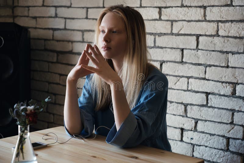 Het blonde meisje zit met hoofdtelefoons en kijkt zorgvuldig in de afstand, die voor haar vingers combineren stock fotografie Het blonde meisje zit met hoofdtelefoons en kijkt zorgvuldig in de afstand, die voor haar vingers combineren stock fotografie