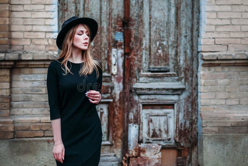 Het blondemeisje met lang haar, in zwarte kleding in hoed, bevindt zich op de achtergrond van het uitstekende antieke oude houten royalty-vrije stock fotografie Het blondemeisje met lang haar, in zwarte kleding in hoed, bevindt zich op de achtergrond van het uitstekende antieke oude houten royalty-vrije stock fotografie