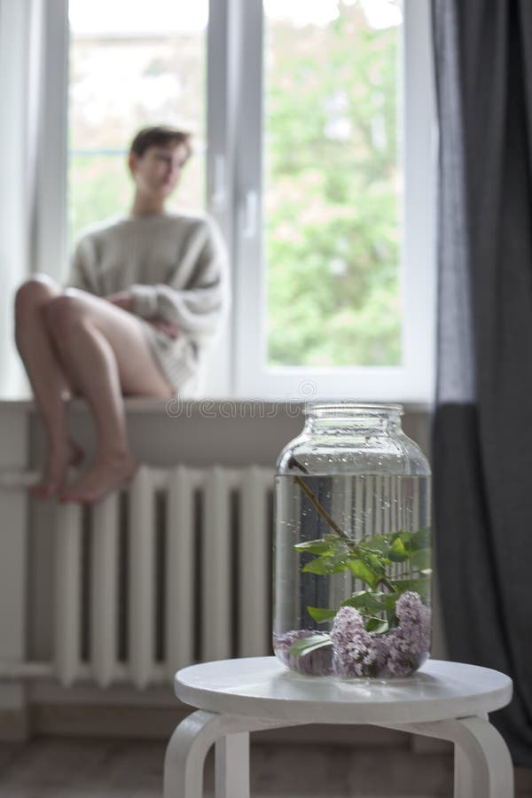 Het boeket van lilac takjes in een transparante kruik op witte stoel als decoratie van binnenland Het meisje zit op het venster royalty-vrije stock fotografie Het boeket van lilac takjes in een transparante kruik op witte stoel als decoratie van binnenland Het meisje zit op het venster royalty-vrije stock fotografie