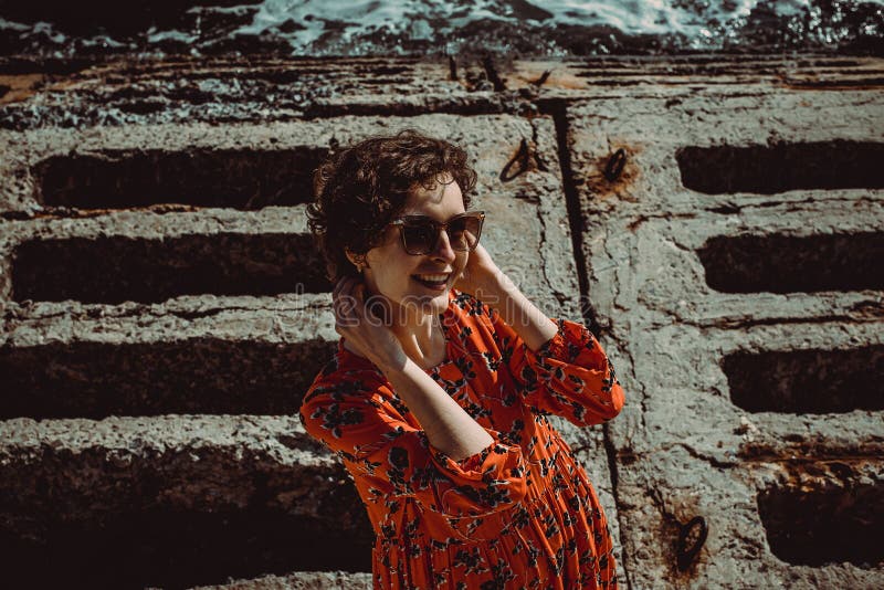 Het charmeren van jonge glimlachende vrouw die in rode kleding met kort krullend haar camera bekijkt De vrouwelijke retro zonnebr stock fotografie Het charmeren van jonge glimlachende vrouw die in rode kleding met kort krullend haar camera bekijkt De vrouwelijke retro zonnebr stock fotografie