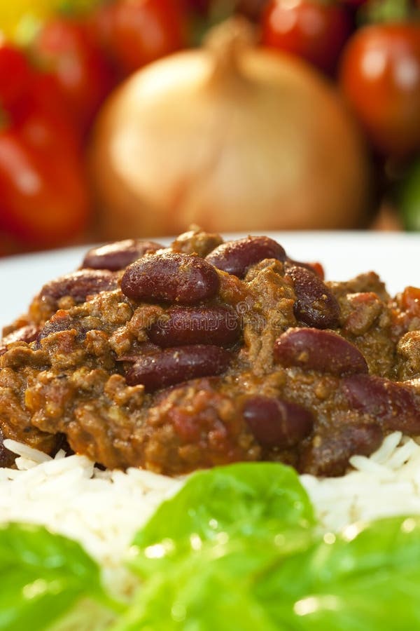 Chili Con Carne on white rice with basil garnish and onions and tomatoes out of focus in the background. Chili Con Carne on white rice with basil garnish and onions and tomatoes out of focus in the background