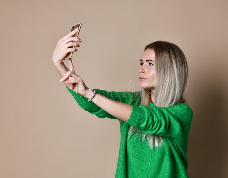Het close-upportret van de jonge vrolijke vrouw van het manierblonde in sweaterslijtage maakt selfie op smartphone, over beige ac stock foto Het close-upportret van de jonge vrolijke vrouw van het manierblonde in sweaterslijtage maakt selfie op smartphone, over beige ac stock foto