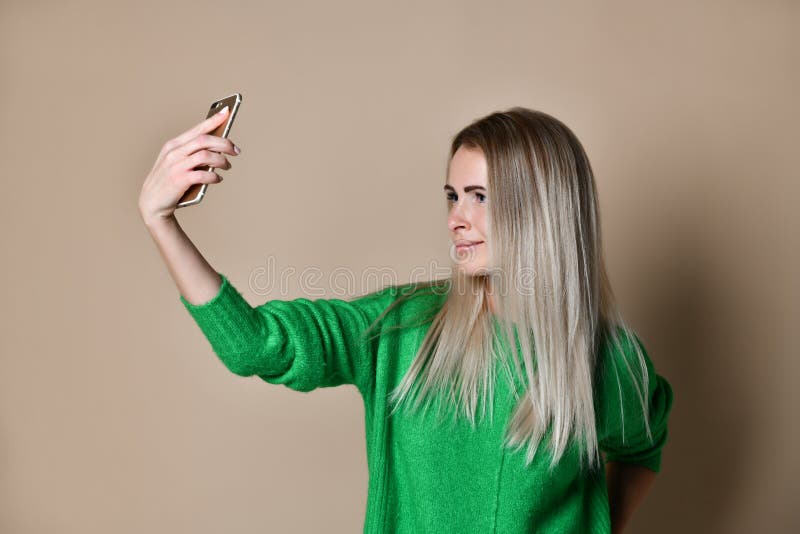 Het close-upportret van de jonge vrolijke vrouw van het manierblonde in sweaterslijtage maakt selfie op smartphone, over beige ac stock afbeeldingen Het close-upportret van de jonge vrolijke vrouw van het manierblonde in sweaterslijtage maakt selfie op smartphone, over beige ac stock afbeeldingen
