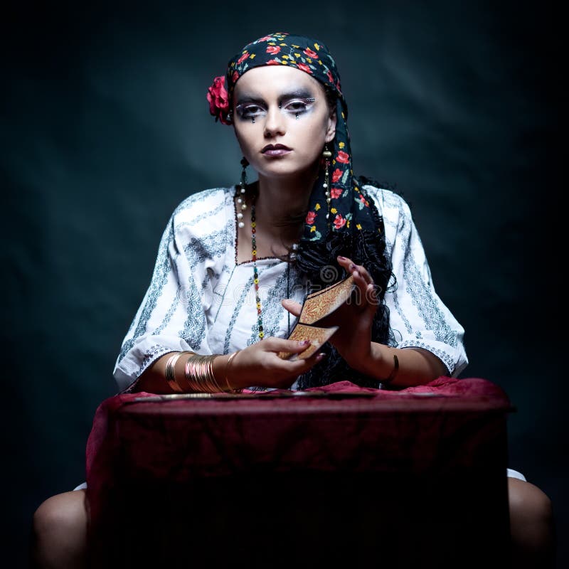 A portrait of a gypsy fortune teller, sitting at a table and mixing the tarot cards that she holds in her hands. she is looking at the camera. A portrait of a gypsy fortune teller, sitting at a table and mixing the tarot cards that she holds in her hands. she is looking at the camera.