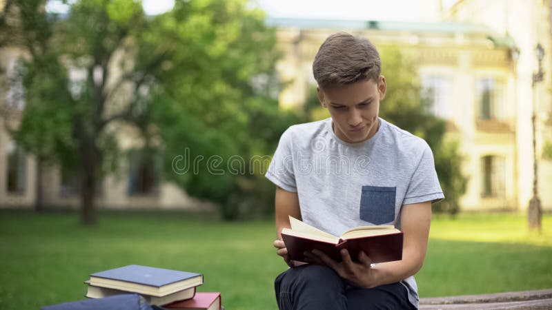 Het geconcentreerde boek van de tienerlezing in park op bank, die voor examens voorbereidingen treffen stock fotografie Het geconcentreerde boek van de tienerlezing in park op bank, die voor examens voorbereidingen treffen stock fotografie
