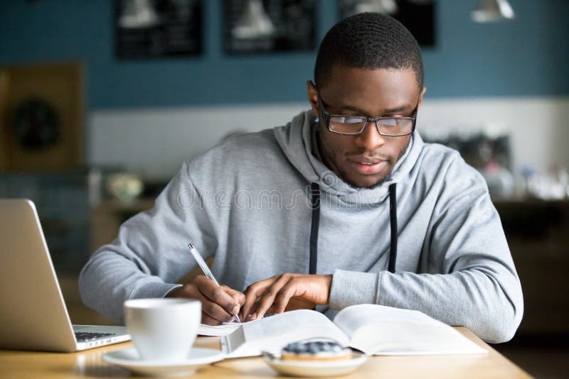 Focused millennial african american student in glasses making notes writing down information from book in cafe preparing for test or exam, young serious black man studying or working in coffee house. Focused millennial african american student in glasses making notes writing down information from book in cafe preparing for test or exam, young serious black man studying or working in coffee house