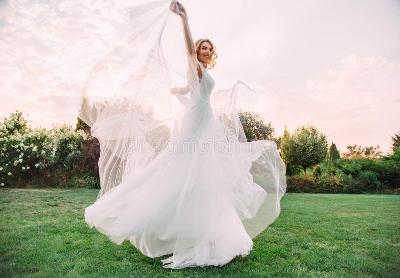 Het gelukkige jonge blondemeisje in een elegante verbazende lange witte huwelijks lichte en vliegende kleding wervelt en glimlach stock afbeeldingen Het gelukkige jonge blondemeisje in een elegante verbazende lange witte huwelijks lichte en vliegende kleding wervelt en glimlach stock afbeeldingen