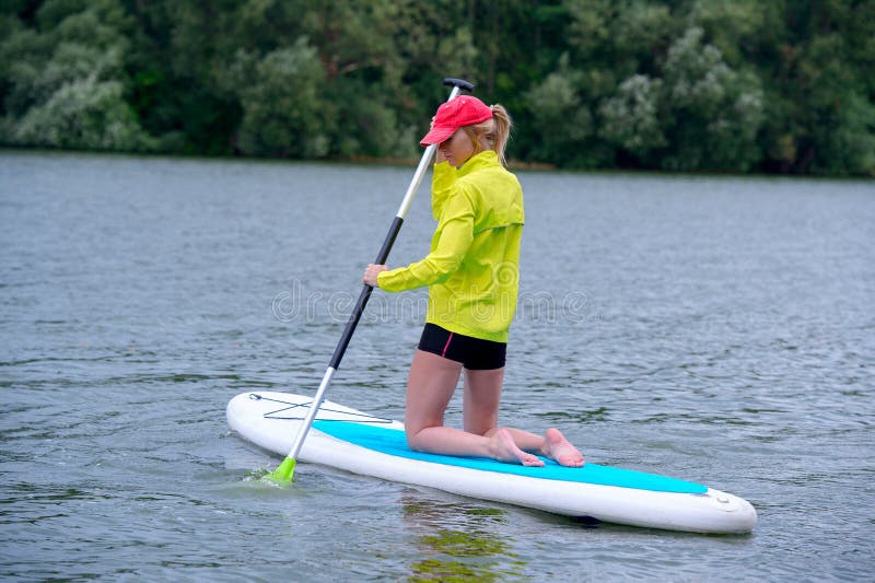 Het gelukkige meisje in een jasje knielt op een surfplank en houdt een lange peddel Achtermeningsfoto royalty-vrije stock fotografie Het gelukkige meisje in een jasje knielt op een surfplank en houdt een lange peddel Achtermeningsfoto royalty-vrije stock fotografie