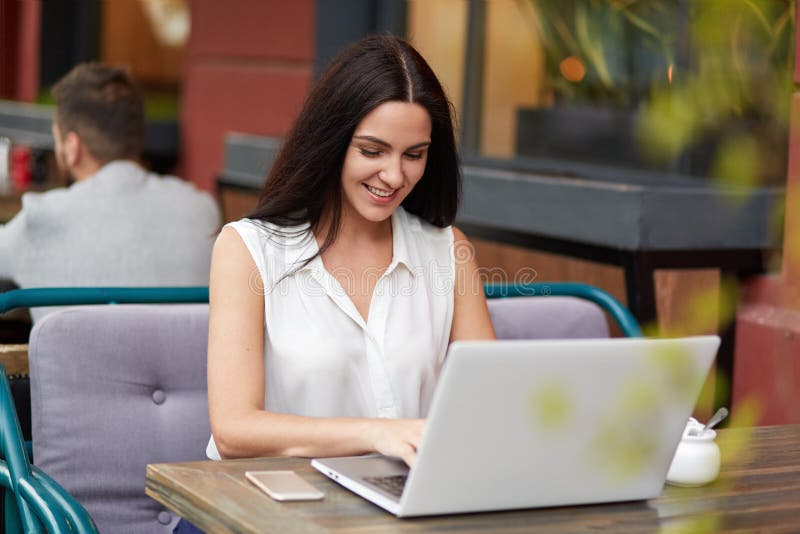 Het gelukkige schitterende jonge wijfje met lang recht donker haar, heeft prettige glimlach, intikt iets op laptop computer, binn royalty-vrije stock afbeelding Het gelukkige schitterende jonge wijfje met lang recht donker haar, heeft prettige glimlach, intikt iets op laptop computer, binn royalty-vrije stock afbeelding