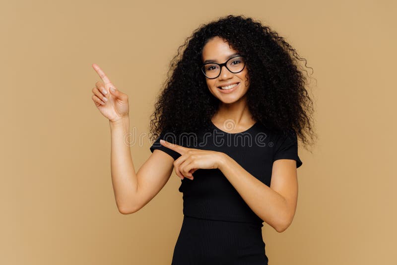 Isolated shot of happy African American woman points up and aside, demonstrates blank copy space for your advertisement, wears spectacles, casual t shirt, has gentle smile on face. People and promo. Isolated shot of happy African American woman points up and aside, demonstrates blank copy space for your advertisement, wears spectacles, casual t shirt, has gentle smile on face. People and promo