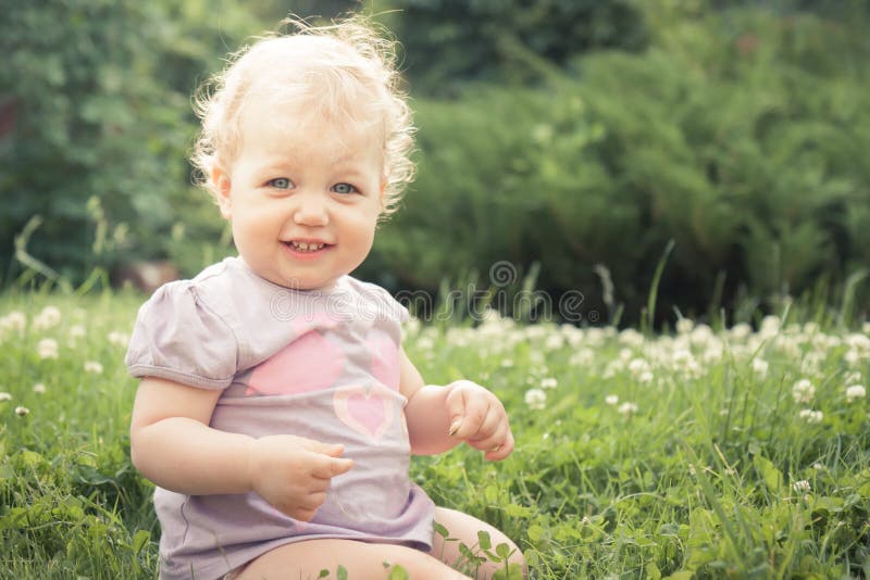Het glimlachen de zitting van het babymeisje in gras onder het tot bloei komen bloeit in de zomerpark in zonnige dag met exemplaa stock afbeeldingen Het glimlachen de zitting van het babymeisje in gras onder het tot bloei komen bloeit in de zomerpark in zonnige dag met exemplaa stock afbeeldingen