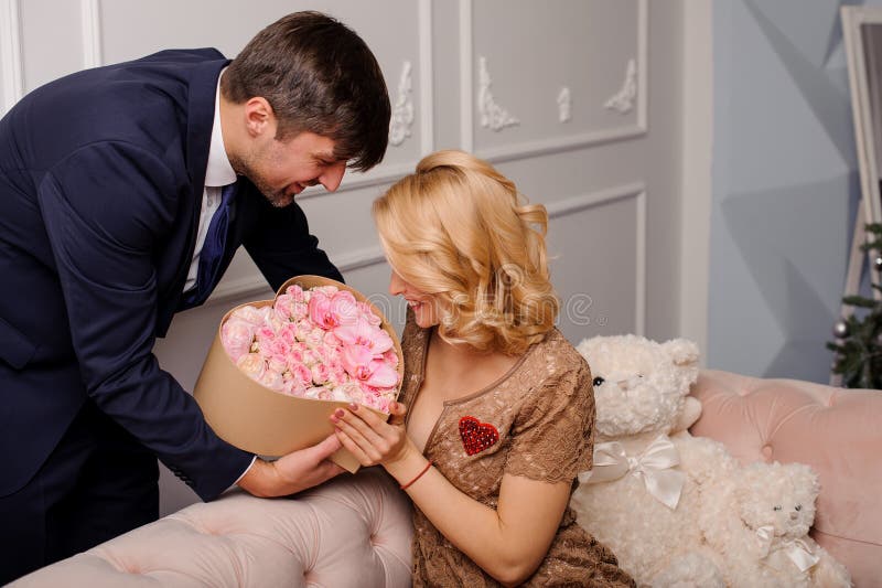 Het glimlachende meisje in de beige kledingszitting op de laag krijgt een doos van bloemen van de haar mens royalty-vrije stock foto Het glimlachende meisje in de beige kledingszitting op de laag krijgt een doos van bloemen van de haar mens royalty-vrije stock foto