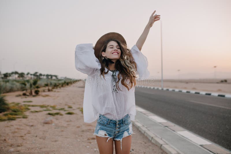 Smiling cheerful long-haired girl with curly hair breathes a full breast and enjoys freedom, standing next to road. Portrait of adorable young woman in white blouse and denim shorts having fun outside. Smiling cheerful long-haired girl with curly hair breathes a full breast and enjoys freedom, standing next to road. Portrait of adorable young woman in white blouse and denim shorts having fun outside