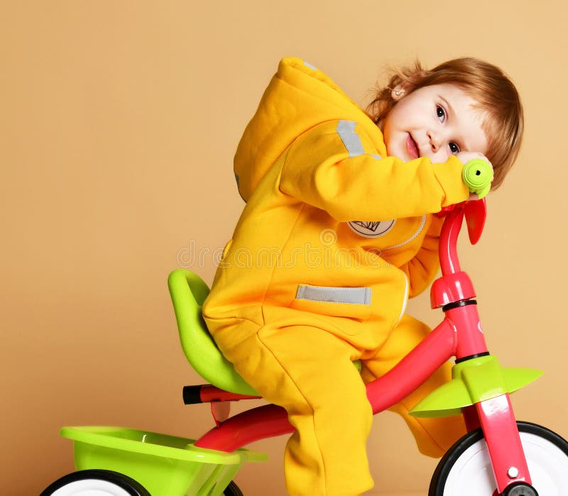 Het jonge geitje die van het babymeisje haar eerste fietsdriewieler in warme gele overall berijden die omhoog op lichtgrijs eruit royalty-vrije stock afbeelding Het jonge geitje die van het babymeisje haar eerste fietsdriewieler in warme gele overall berijden die omhoog op lichtgrijs eruit royalty-vrije stock afbeelding