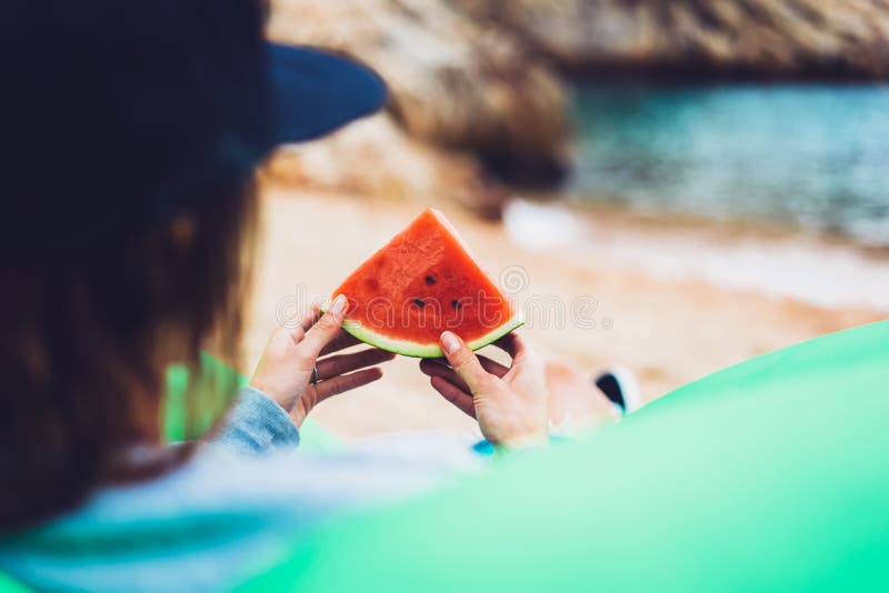 Het jonge meisje hipster ontspant op strandkust en houdt in haar hand een plak van rode vers fruitwatermeloen op blauwe overzeese royalty-vrije stock afbeelding Het jonge meisje hipster ontspant op strandkust en houdt in haar hand een plak van rode vers fruitwatermeloen op blauwe overzeese royalty-vrije stock afbeelding