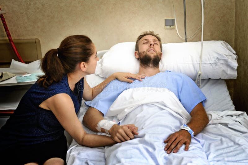 Het jonge paar bij de mens die van de het ziekenhuisruimte in bed liggen maakte zich vrouw ongerust die zijn hand het geven houde royalty-vrije stock afbeelding Het jonge paar bij de mens die van de het ziekenhuisruimte in bed liggen maakte zich vrouw ongerust die zijn hand het geven houde royalty-vrije stock afbeelding