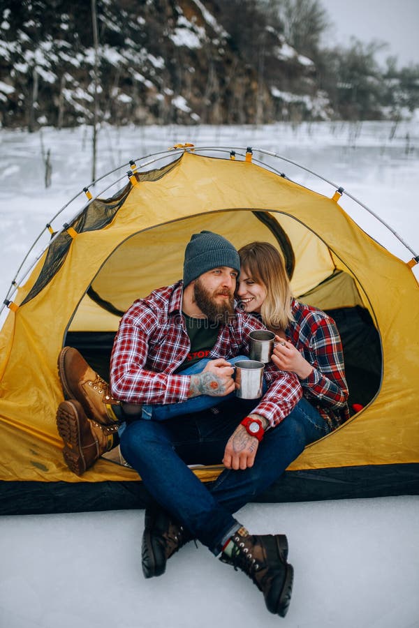 Young couple sits in yellow tourist tent and drinks hot tea from mugs during winter hike. Young couple sits in yellow tourist tent and drinks hot tea from mugs during winter hike.