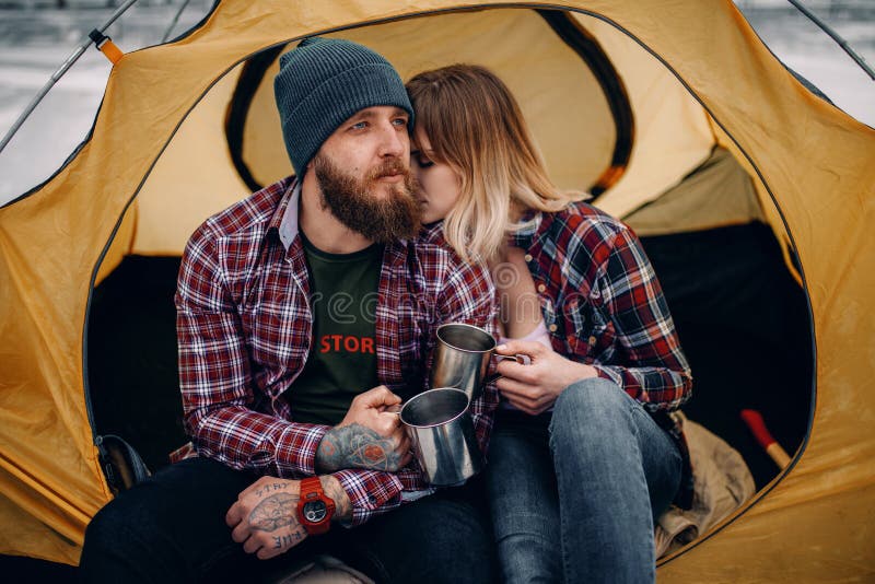 Young couple sits in yellow tourist tent and drinks hot tea from mugs during winter hike. Young couple sits in yellow tourist tent and drinks hot tea from mugs during winter hike.