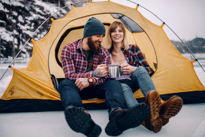Young couple sits in yellow tourist tent, smiles and drinks hot tea from mugs during winter hike. Young couple sits in yellow tourist tent, smiles and drinks hot tea from mugs during winter hike.