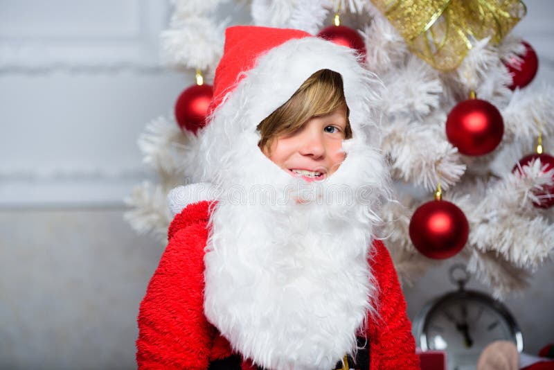 Het jongensjonge geitje kleedde zich als santa met witte kunstmatige baard en rode hoed dichtbij Kerstmisboom Het kostuum van de royalty-vrije stock fotografie Het jongensjonge geitje kleedde zich als santa met witte kunstmatige baard en rode hoed dichtbij Kerstmisboom Het kostuum van de royalty-vrije stock fotografie