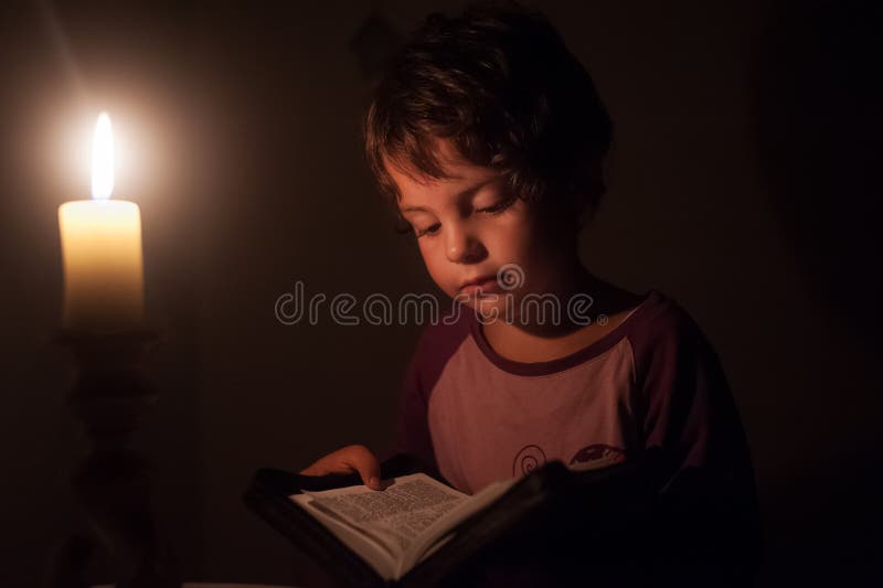 Het kind leest het boek in de donkere kamer door het licht van een waskaars royalty-vrije stock afbeeldingen Het kind leest het boek in de donkere kamer door het licht van een waskaars royalty-vrije stock afbeeldingen