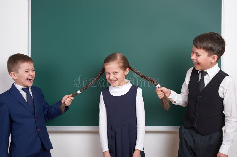 Het kindspel en het hebben van pret, jongens trekken de meisjesvlechten, dichtbij de lege achtergrond van het schoolbord, gekleed royalty-vrije stock afbeeldingen Het kindspel en het hebben van pret, jongens trekken de meisjesvlechten, dichtbij de lege achtergrond van het schoolbord, gekleed royalty-vrije stock afbeeldingen